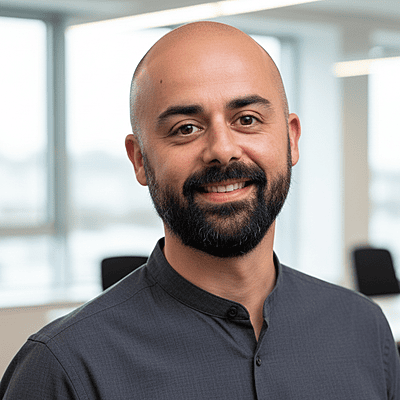 Photo of a Spanish man with a bald head, black coloured full beard and mustache wearing a grey collarless shirt.