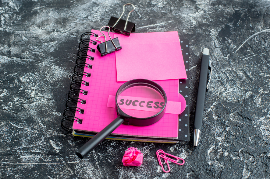 A notebook with a magnifying glass looking at at the word success and pens and paper clips around them