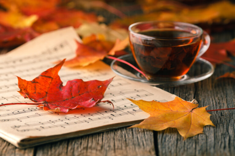 Photo of a wooden table with a music score and a glass cup of tea beside it, with autumnal coloured leaves scattered around.