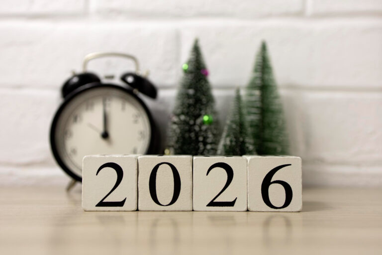 Photo of some wooden blocks showing '2026' with an alarm clock and some christmas trees in the backgroun against a white painted brick wall.