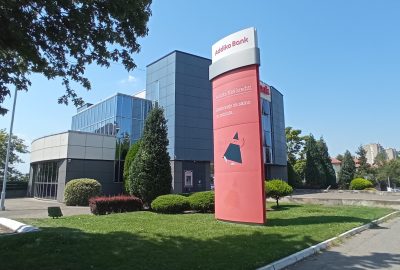 Photo of Addiko Bank - a large glass building with a tall red sign in front and lush green landscaping.