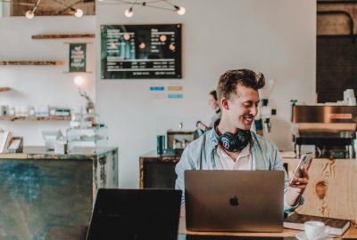 Photo of a man in a coffee shop with a laptop looking at a mobile phone