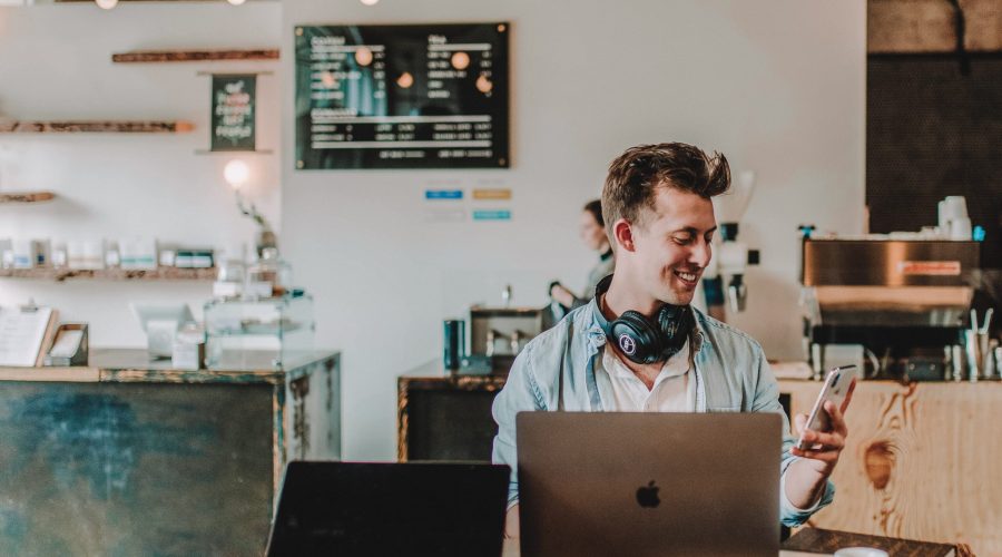 Photo of a man in a coffee shop with a laptop looking at a mobile phone